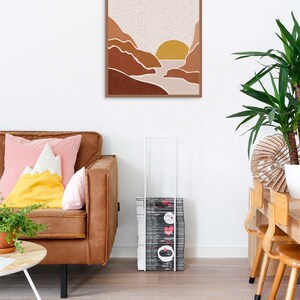 May include: A framed print of a minimalist landscape with a sunset over a river between two mountains. The mountains are brown and the sky is beige with white dots. The sun is yellow.