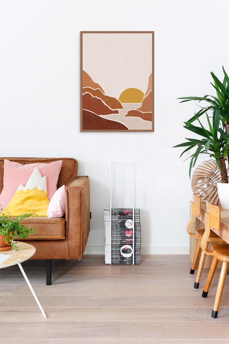 May include: A framed print of a minimalist landscape with a sunset over a river between two mountains. The mountains are brown and the sky is beige with white dots. The sun is yellow.