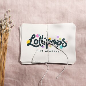 May include: A stack of white cards tied with string, featuring the text "Lollipops KIDS ACADEMY" in a playful font with colorful accents. The cards rest on a pink textured surface, with dried flowers to the left.