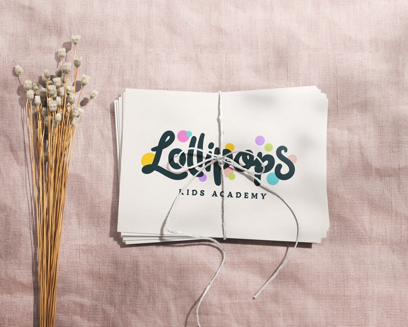 May include: A stack of white cards tied with string, featuring the text "Lollipops KIDS ACADEMY" in a playful font with colorful accents. The cards rest on a pink textured surface, with dried flowers to the left.