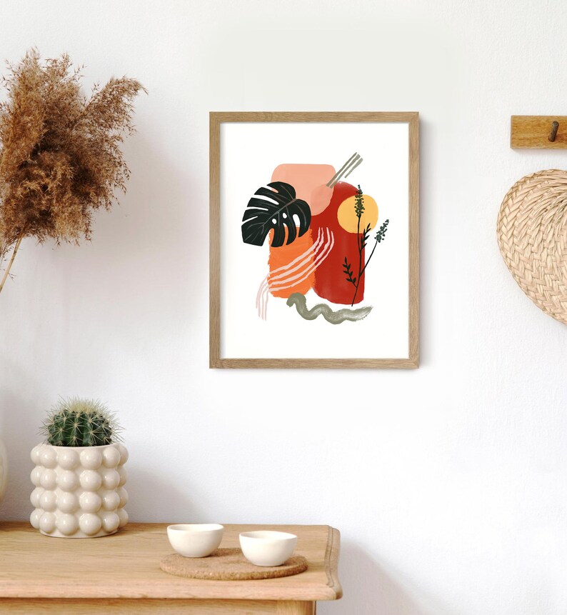 May include: Abstract art print with a red, orange, and pink color scheme. The print features a large leaf, a sun, and a few lines. The print is framed in a light wood frame.