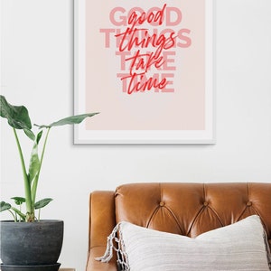 May include: A framed print with the words "Good Things Take Time" in red and pink lettering. The print hangs above a brown leather couch with a decorative pillow. A potted plant sits on a wooden side table.