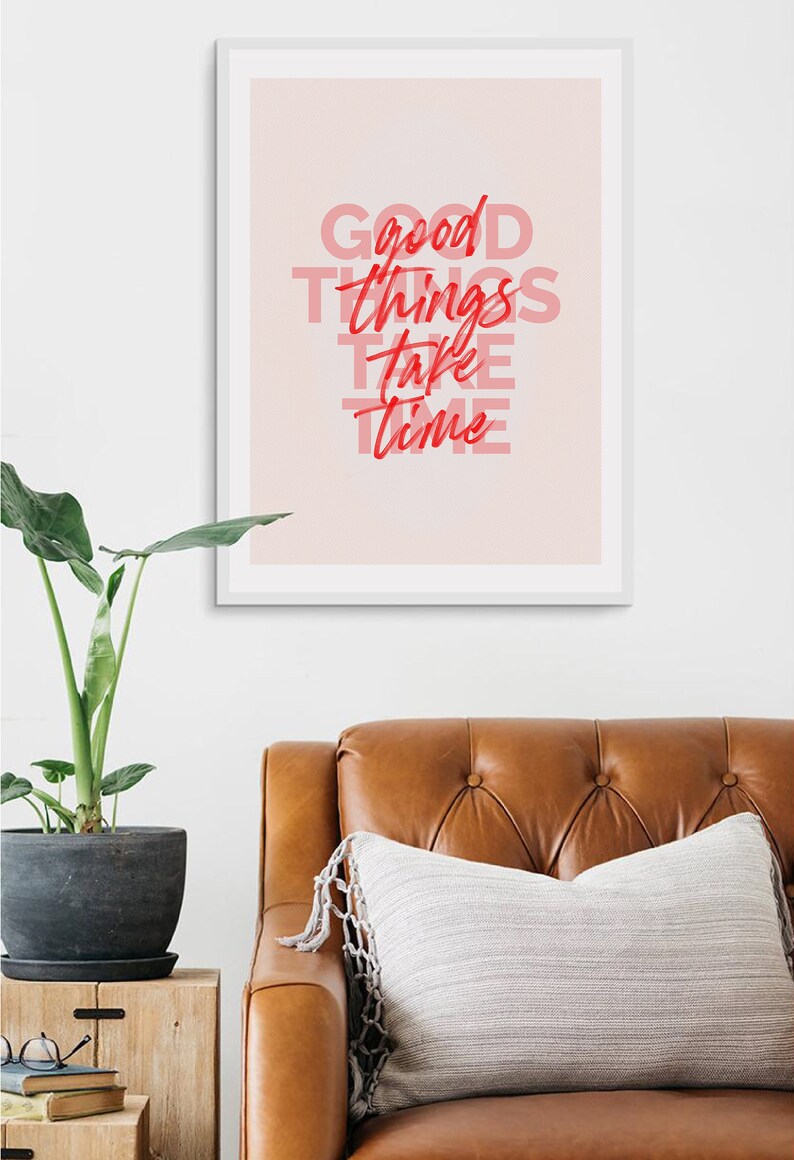 May include: A framed print with the words "Good Things Take Time" in red and pink lettering. The print hangs above a brown leather couch with a decorative pillow. A potted plant sits on a wooden side table.