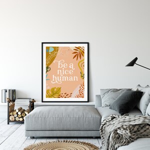May include: A framed print with a peach background and abstract shapes in shades of brown, orange, and green. The text "be a nice human" is written in white cursive font.