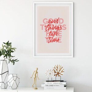 May include: A framed print with the text "Good things take time" in red and pink. The print is displayed on a white wall above a white cabinet. Decorative items include vases, a wooden figure, and sculptures.