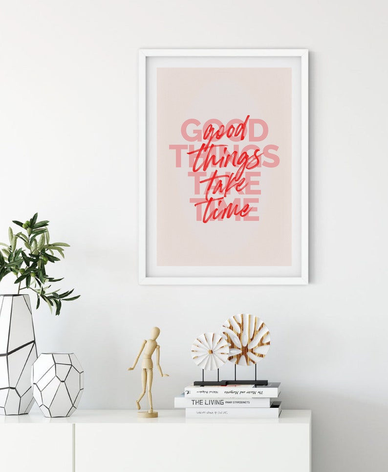 May include: A framed print with the text "Good things take time" in red and pink. The print is displayed on a white wall above a white cabinet. Decorative items include vases, a wooden figure, and sculptures.