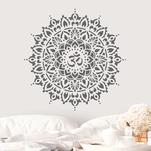 May include: Gray mandala stencil with an Om symbol in the center. The stencil is on a white wall with a bed and a vase of white flowers in the background.