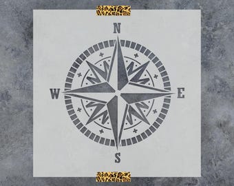 May include: A square stencil featuring a dark gray compass rose design with cardinal directions N, S, E, and W. The compass has a central star shape surrounded by a circular border with small plus signs. Two leopard print tape strips are attached.