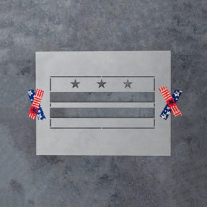 May include: A rectangular light gray stencil featuring a cutout design of the Washington D.C. flag, which includes two horizontal bars and three five-pointed stars centered in the top section. Two pieces of patterned tape are attached to the sides.