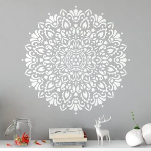 May include: A large white circular mandala wall decal featuring intricate symmetrical floral and leaf patterns. The design is displayed on a solid gray wall above a stack of books, a white deer figurine, and a glass jar filled with red chili peppers.