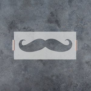 May include: A white stencil with a black mustache shape cut out. The stencil is taped to a gray background.