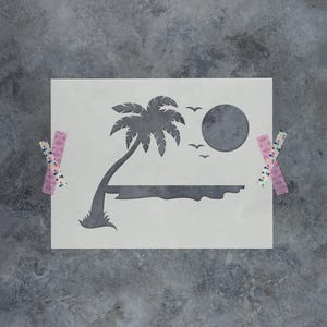 May include: A rectangular stencil sheet featuring a cutout design of a palm tree, a large circular sun, and three small birds flying above a horizontal line representing the horizon. The stencil is secured with patterned adhesive tape.