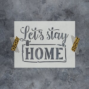 May include: White stencil with the text "Let's stay HOME" in a black, handwritten font. The stencil is taped to a gray, textured surface with leopard print tape.