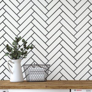 May include: A white ceramic pitcher with a handle, a black wire basket with white towels inside, and a green leafy plant in the pitcher. The background is a white wall with a black herringbone pattern.