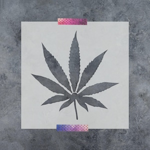 May include: A square stencil featuring a dark gray, seven-pointed cannabis leaf cutout in the center. The stencil is secured at the top and bottom edges with two strips of purple and pink gradient washi tape with a subtle dot pattern.