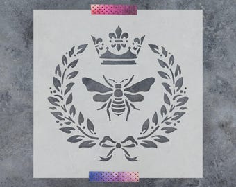 May include: A square stencil featuring a central bee icon surrounded by a circular laurel wreath. Above the bee is a crown, and below the wreath is a decorative bow. The stencil is light gray and is secured with two strips of patterned tape.