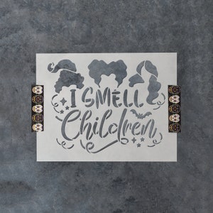 May include: A white stencil with the text "I smell children" and a bat graphic. Three witch silhouettes are above the text. The stencil is on a gray background.