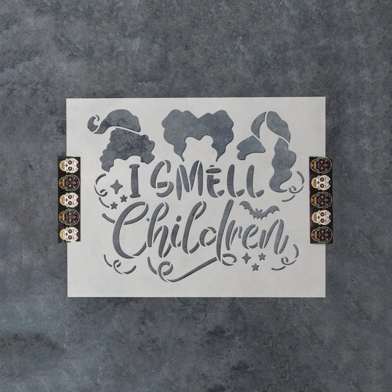 May include: A white stencil with the text "I smell children" and a bat graphic. Three witch silhouettes are above the text. The stencil is on a gray background.