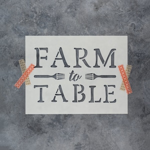 May include: A white stencil with the text "FARM to TABLE" in black lettering. The stencil is on a gray background with two pieces of orange and white patterned tape on either side.