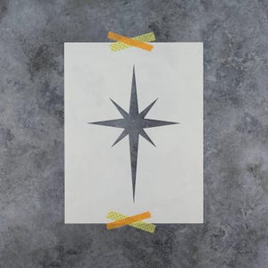 May include: A rectangular stencil sheet featuring a centered, eight-pointed star design with elongated vertical points. The stencil is secured at the top and bottom edges with two pieces of patterned, orange and green adhesive tape.
