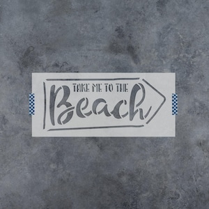 May include: A rectangular stencil featuring a directional arrow shape and the text "TAKE ME TO THE BEACH" in a combination of block and script lettering. The stencil is light gray and includes small blue patterned tape tabs on the left and right sides.
