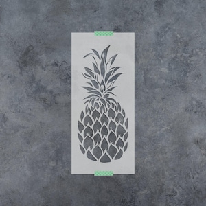 May include: A rectangular stencil featuring a cutout design of a pineapple with a detailed leafy top and a textured fruit body. The stencil is semi-transparent, light gray, and secured at the top and bottom edges with small pieces of green tape.