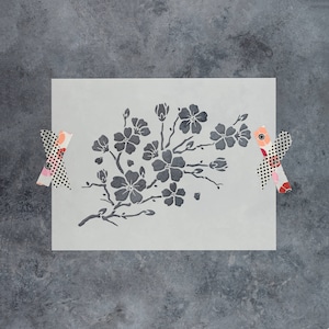 May include: A black and white stencil of a floral design featuring a branch with several blossoms. The stencil is on a light gray background.