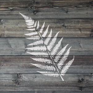 May include: A white fern frond stencil on a weathered grey wood background.