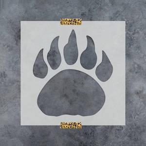 May include: A square stencil featuring a large bear paw print cutout in the center. The stencil is semi-transparent and is secured at the top and bottom edges with two strips of adhesive tape that have a brown and black leopard print pattern.
