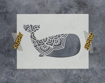 May include: A rectangular white stencil featuring a cutout of a whale silhouette. The whale's tail and rear body are decorated with an intricate, symmetrical mandala-style pattern. The stencil is secured at the sides with pieces of patterned tape.