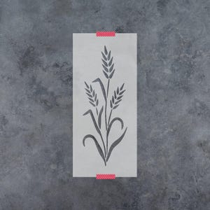 May include: A rectangular stencil featuring a cutout design of three stalks of wheat with leaves. The stencil is semi-transparent and off-white, secured at the top and bottom edges with small strips of red and white polka-dot patterned tape.