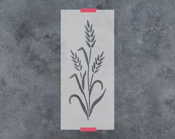 May include: A rectangular stencil featuring a cutout design of three stalks of wheat with leaves. The stencil is semi-transparent and off-white, secured at the top and bottom edges with small strips of red and white polka-dot patterned tape.