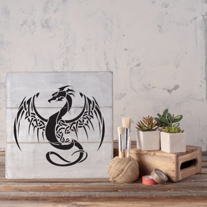 K&ouml;nnte beinhalten: Ein Holzschild mit einem schwarzen und wei&szlig;en Tribal-Drachen-Design. Das Schild steht auf einem Holztisch mit einer Schnurkugel, Pinseln und Topfpflanzen.