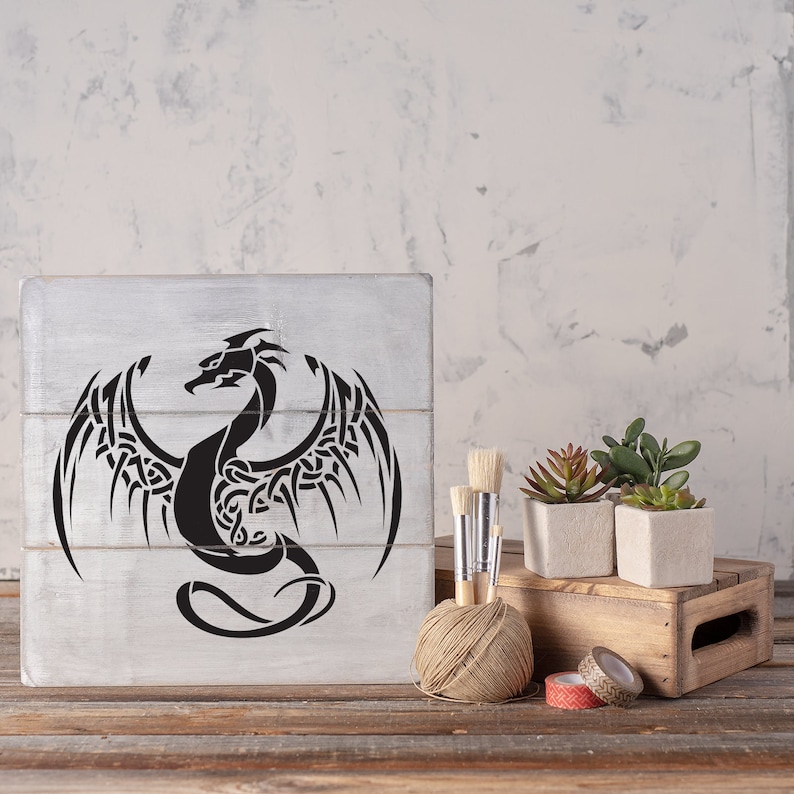 K&ouml;nnte beinhalten: Ein Holzschild mit einem schwarzen und wei&szlig;en Tribal-Drachen-Design. Das Schild steht auf einem Holztisch mit einer Schnurkugel, Pinseln und Topfpflanzen.