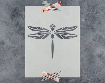 May include: A rectangular stencil sheet featuring a centered, dark gray dragonfly silhouette with detailed wing patterns. The stencil is secured at the top and bottom edges with two pieces of decorative black and white polka dot washi tape.