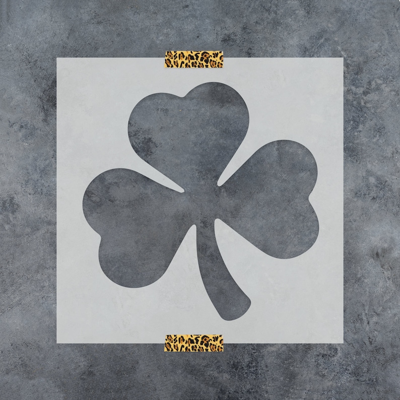 May include: A white stencil of a four-leaf clover, a traditional Irish symbol of good luck. The stencil is taped to a gray textured surface.