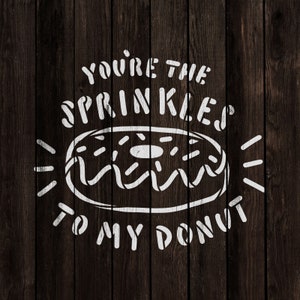 Op de afbeelding: Witte tekst op een donkere houten achtergrond luidt "You're the sprinkles to my donut". Een witte omtrek van een donut met sprenkels bevindt zich in het midden van de tekst.