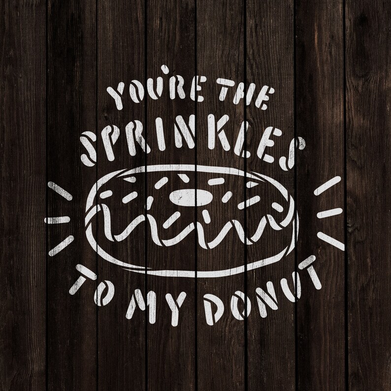 Op de afbeelding: Witte tekst op een donkere houten achtergrond luidt "You're the sprinkles to my donut". Een witte omtrek van een donut met sprenkels bevindt zich in het midden van de tekst.