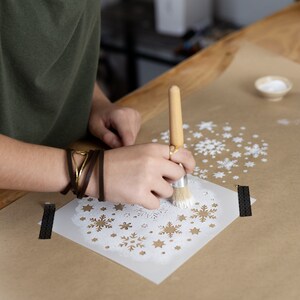 K&ouml;nnte beinhalten: Eine Person verwendet einen Pinsel, um wei&szlig;e Farbe durch eine Schneeflocken-Schablone auf braunes Papier aufzutragen. Die Schablone ist von anderen Schneeflocken-Schablonen umgeben.