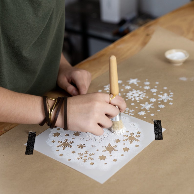 K&ouml;nnte beinhalten: Eine Person verwendet einen Pinsel, um wei&szlig;e Farbe durch eine Schneeflocken-Schablone auf braunes Papier aufzutragen. Die Schablone ist von anderen Schneeflocken-Schablonen umgeben.