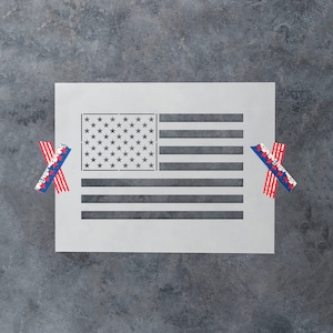 May include: A white stencil of the American flag with red and blue stars and stripes. The stencil is on a gray background and is surrounded by two pieces of red, white, and blue washi tape.