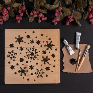 K&ouml;nnte beinhalten: Ein Holzschild mit einem Schneeflocken-Schablonenmuster. Das Schild ist von roten Beeren und gr&uuml;nen Bl&auml;ttern umgeben. Zwei Tuben schwarzer Farbe und zwei Pinsel befinden sich auf einer braunen Papieroberfl&auml;che.