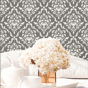 May include: A large bouquet of white hydrangea flowers sits in a clear glass vase. The background features a wall covering with a repeating white damask pattern printed on a dark gray surface, creating a high-contrast decorative visual effect.