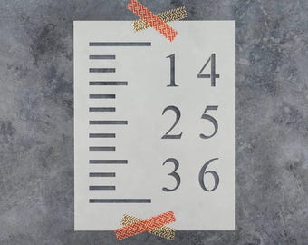 May include: A rectangular white stencil featuring a vertical ruler-style measurement scale on the left side and the numbers 1, 4, 2, 5, 3, and 6 arranged in two columns on the right. The stencil is secured with two pieces of patterned orange tape.