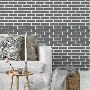 May include: A white sofa with two decorative pillows, one featuring a black and white bird pattern and the other a tan leaf print. A gold side table holds a glass vase with a pink flower, a candle, and a glass votive against a gray brick wall.