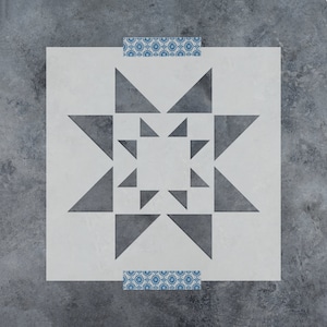 May include: A square stencil with a geometric star design, featuring triangles and a central square. The stencil is white with gray shapes and is taped to a gray surface with blue patterned tape.