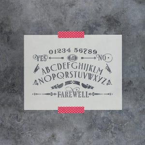 May include: A rectangular stencil featuring a spirit board design with the numbers 0-9, the alphabet, the words YES, NO, and FAREWELL, along with illustrations of pointing hands, arrows, and an eye symbol, secured by two strips of pink polka dot tape.