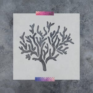 May include: A square, light gray stencil featuring a dark gray, intricate coral reef design in the center. The stencil is secured at the top and bottom edges with two strips of purple and pink patterned adhesive tape.