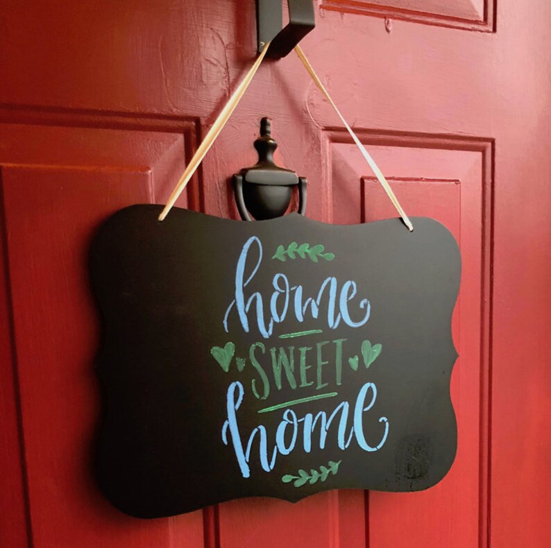 May include: A black chalkboard sign with a white ribbon hanging from a hook on a red door. The sign says "home sweet home" in blue and green chalk lettering with green leaf accents.