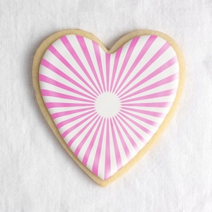 May include: A heart-shaped cookie decorated with pink and white icing in a sunburst pattern.